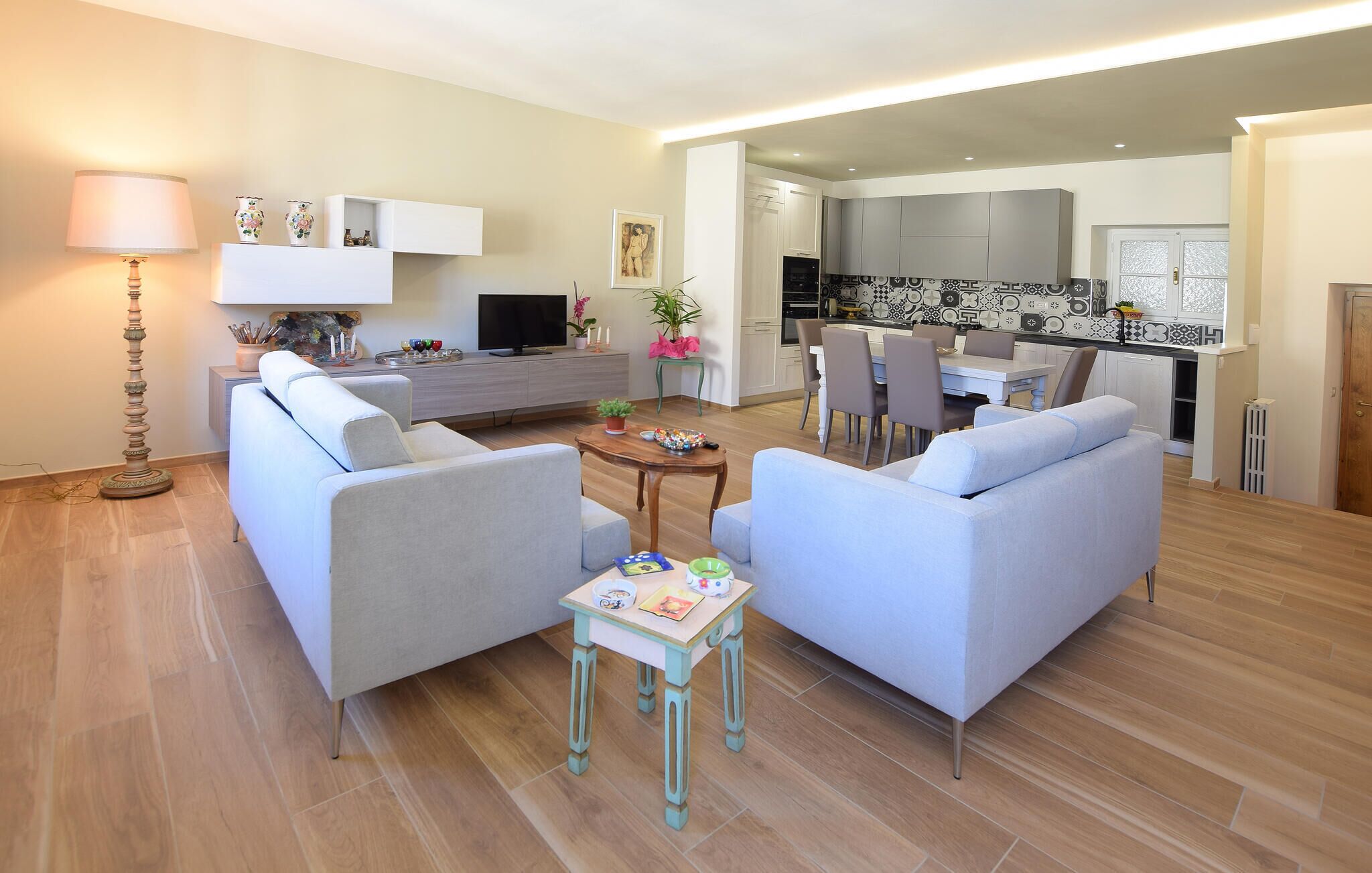 Ein modernes Wohnzimmer mit zwei Sofas, einem Couchtisch und einem Fernseher. Ein Essbereich mit Tisch und Stühlen ist ebenfalls vorhanden. Im Hintergrund ist eine Küche mit modernen Geräten zu sehen.