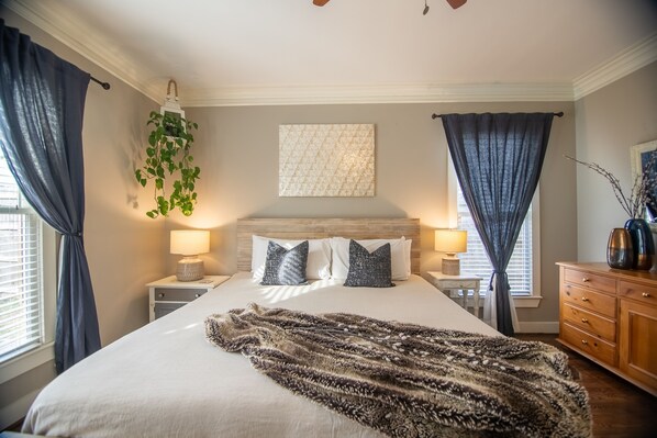 Un dormitorio ordenado con una cama grande, dos mesitas de noche con lámparas, una planta colgante y un aparador de madera.