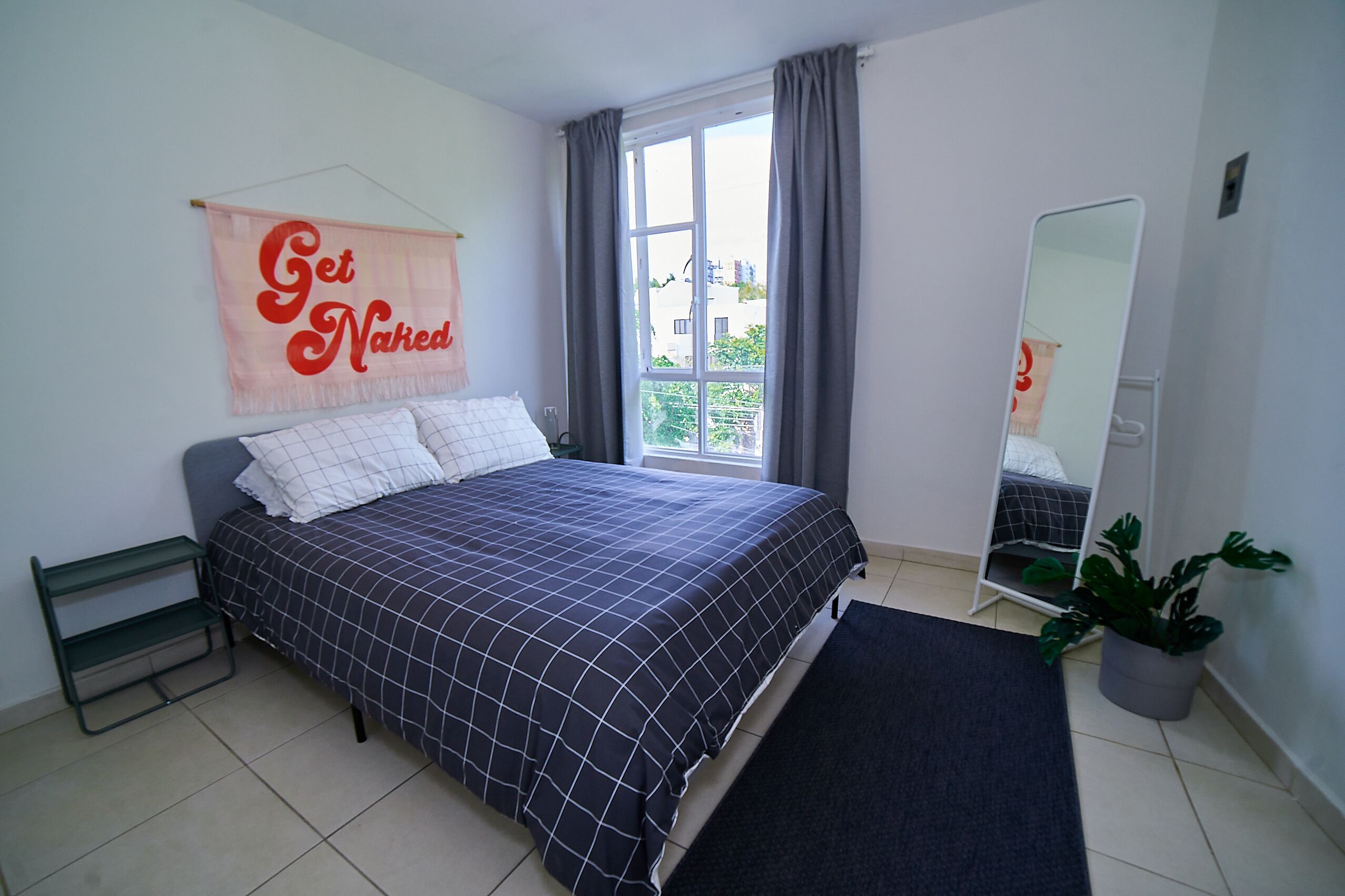 A bedroom with a bed, a bedside table, a mirror, a window with curtains, a potted plant, and a wall with a 'Get Naked' sign.