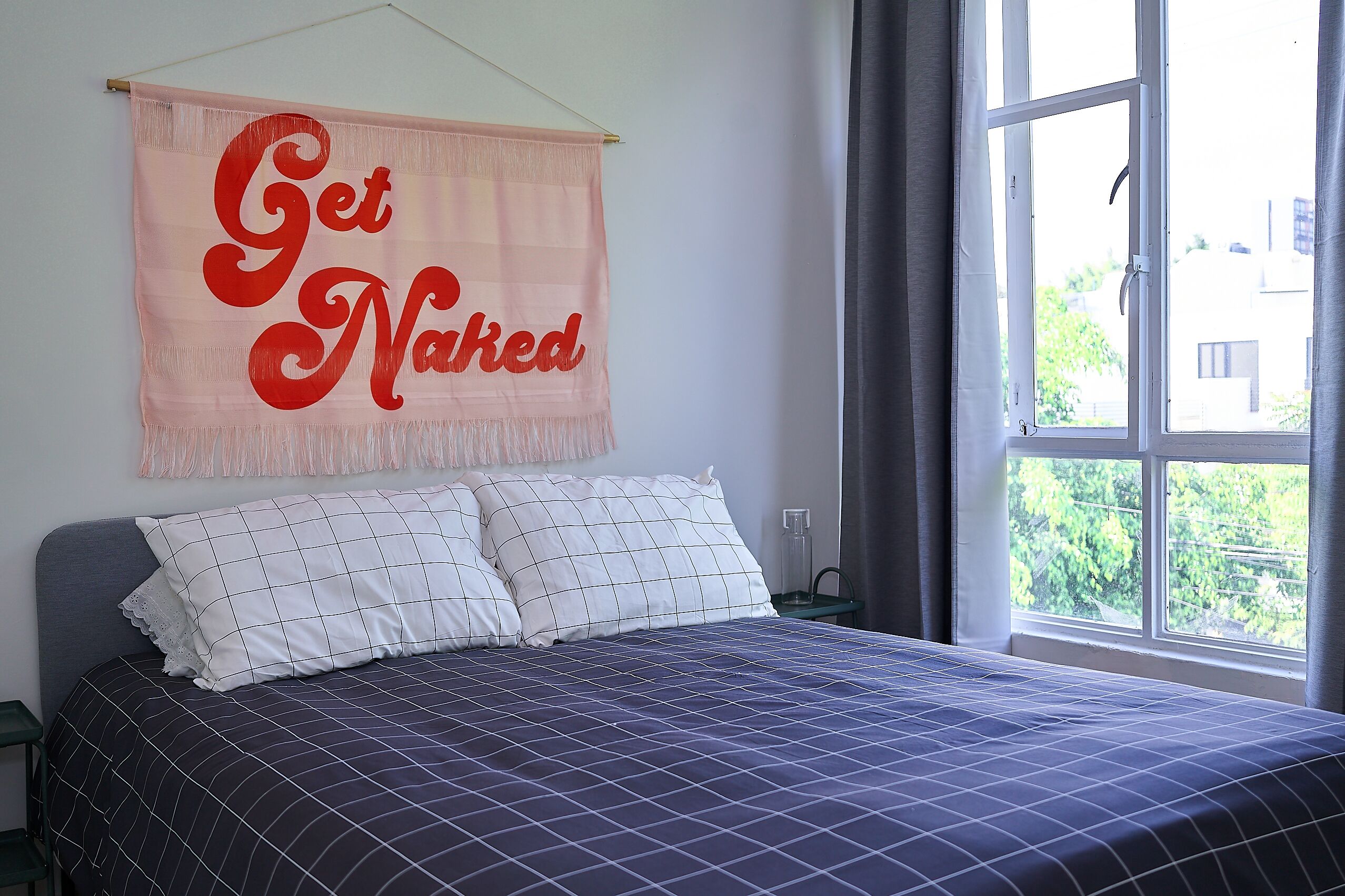 A bedroom with a bed, a window, and a wall hanging that says "Get Naked".