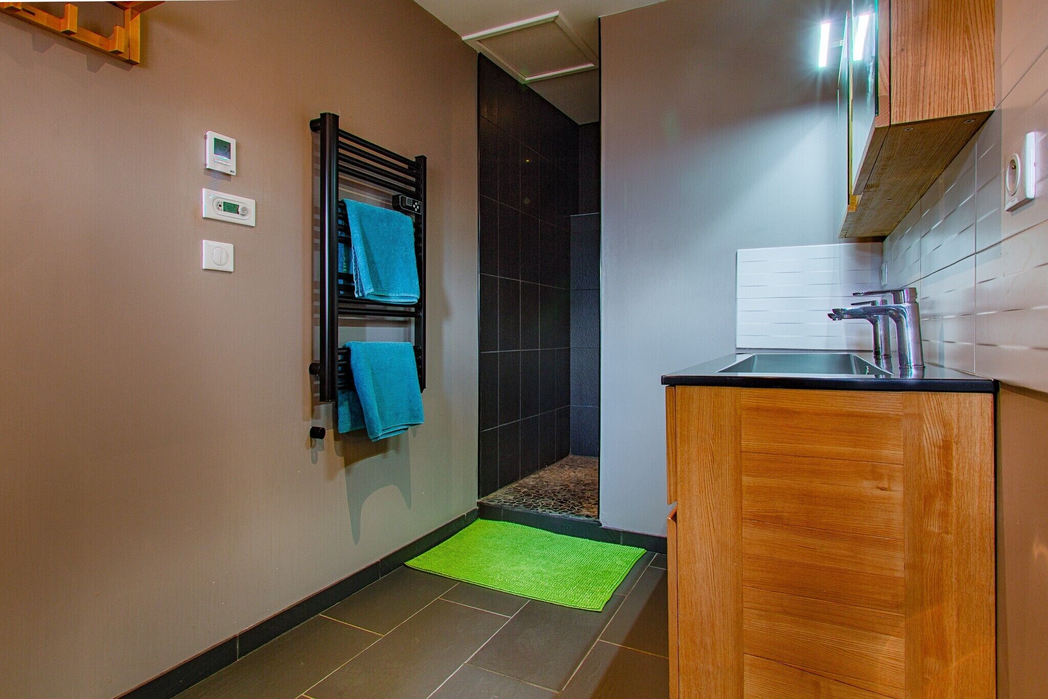 Een badkamer met een douche, een handdoekrek met blauwe handdoeken, een groene mat en een houten kastje.