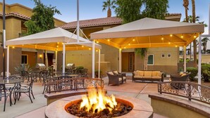 An outdoor patio area with a fire pit, tables, chairs, and umbrellas providing shade.