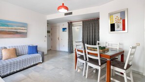 A dining area with a glass table and white chairs, a sofa with patterned cushions, and a painting on the wall. - Apartment "Don Juan Manuel" with Shared Pool & Wi-Fi (Torremolinos)