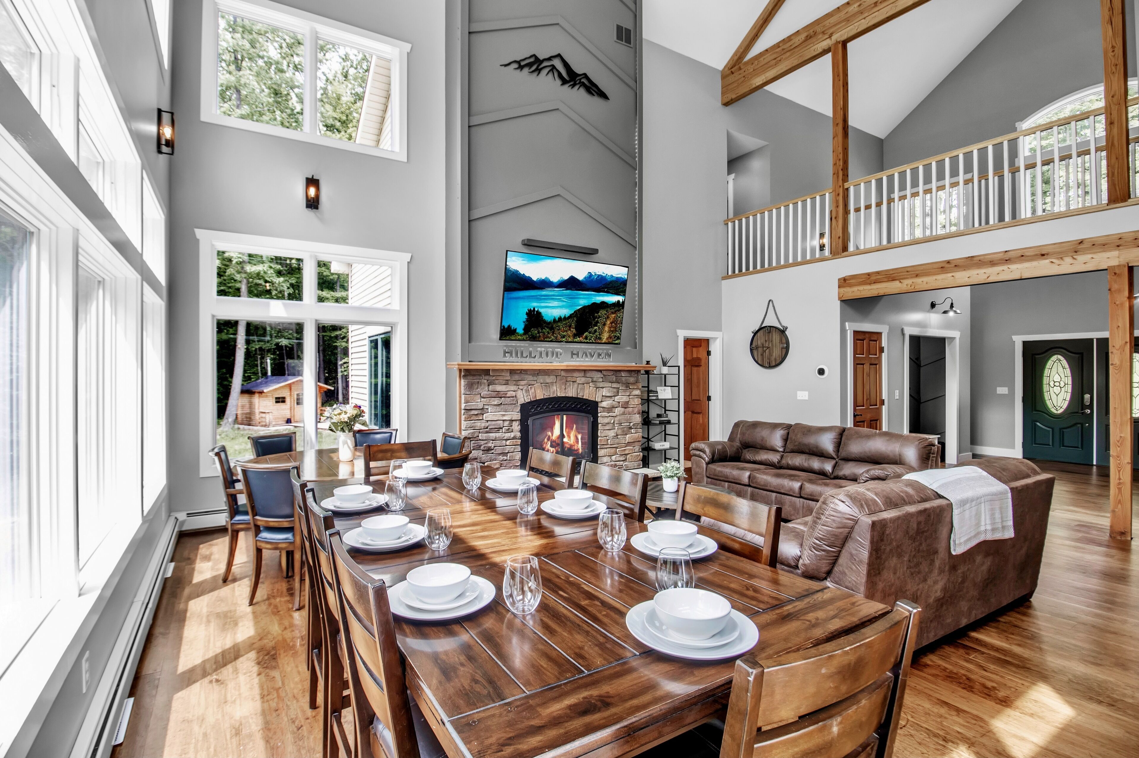 Amplia sala de estar con chimenea, una mesa de comedor lista para seis personas y un gran ventanal con vistas a un paisaje pintoresco.