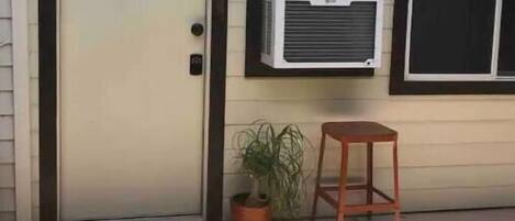Una casa con una puerta cerrada, un equipo de aire acondicionado en la pared, un taburete de madera pequeño y una planta en maceta.