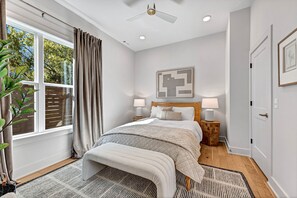 Un dormitorio con cama, lámparas de noche, ventilador de techo, ventana con cortinas e imagen enmarcada en la pared.