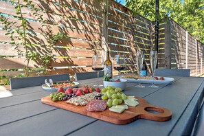 Un espacio exterior para comer con una mesa de madera dispuesta para una comida, incluyendo una tabla de embutidos, copas de vino y una botella de vino.