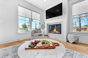 Una sala de estar moderna con chimenea, una mesa redonda de mármol con una tabla de embutidos, un sofá y ventanas amplias.