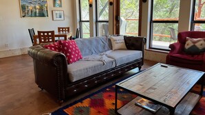 Un salon avec un canapé en cuir, une table basse en bois et un tapis coloré. Il y a deux fenêtres avec des rideaux, une table à manger avec des chaises et des œuvres d’art accrochées au mur.