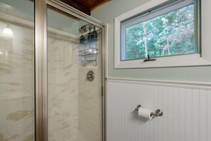 Une salle de bain avec une porte de douche en verre, une fenêtre donnant sur les arbres et un porte-rouleau de papier toilette.