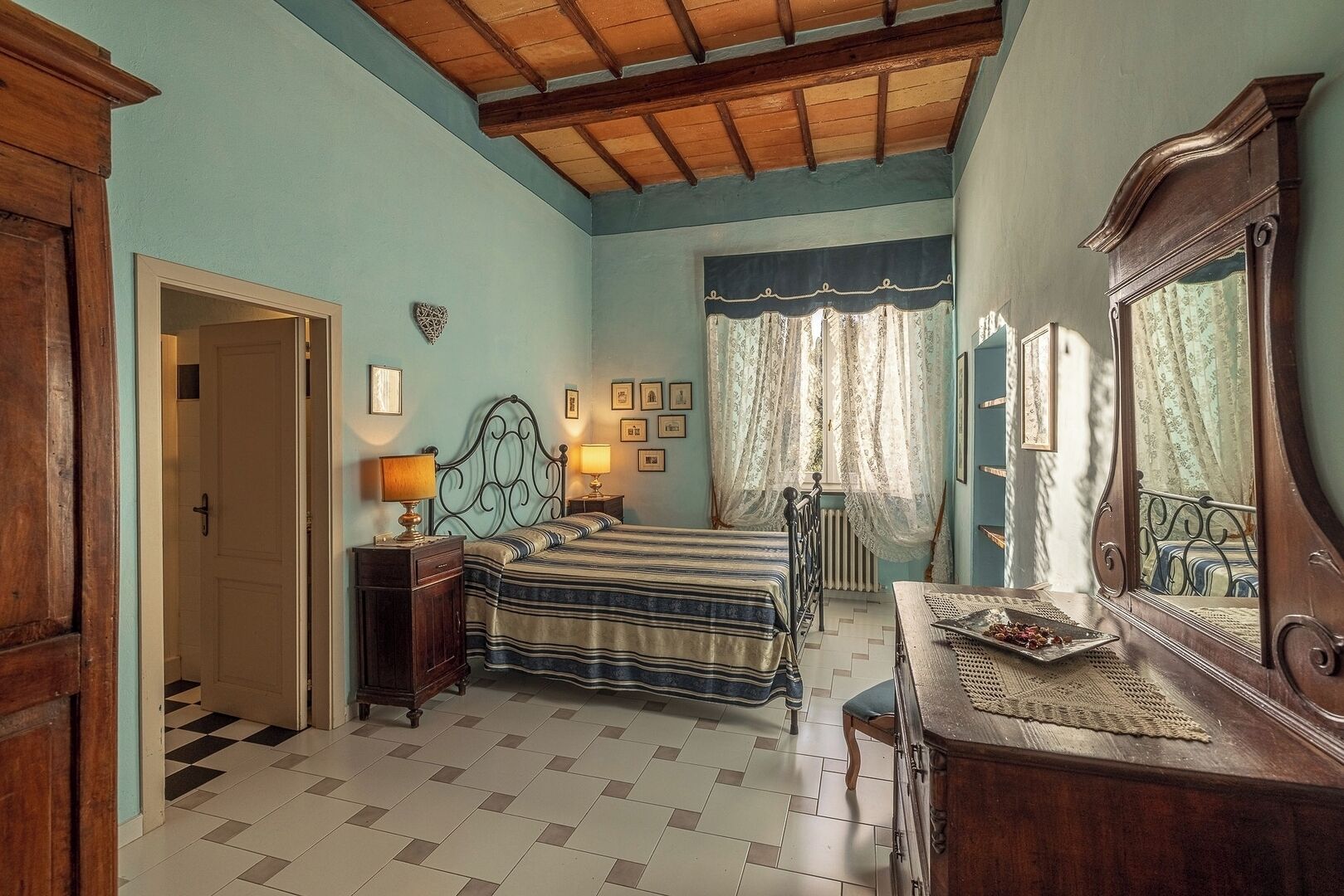 Une chambre à coucher comprenant un lit, des tables de chevet, une commode, un miroir, une fenêtre avec des rideaux et une porte.