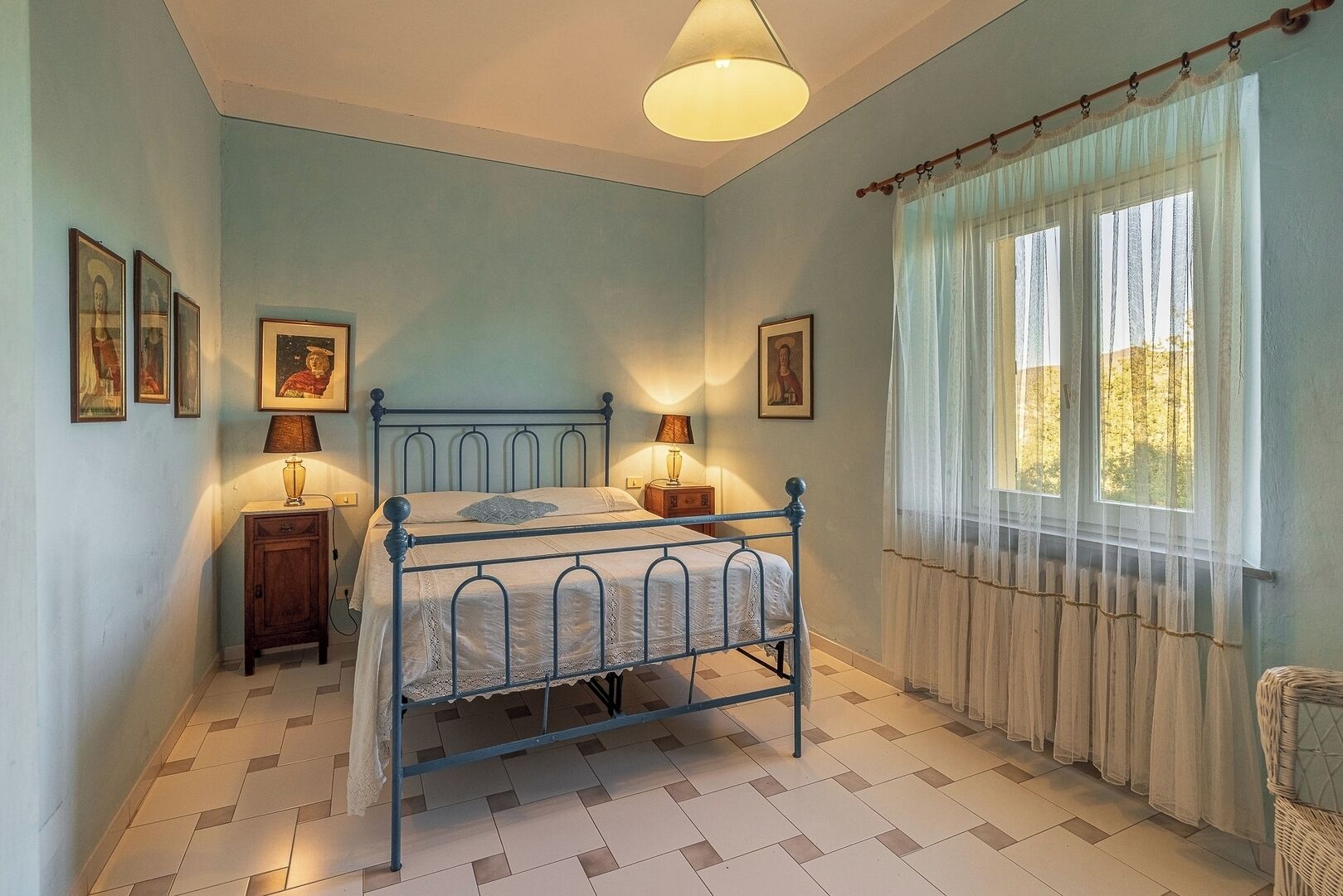 A bedroom with a metal bed frame, two bedside tables with lamps, a window with sheer curtains, and wall-mounted pictures.