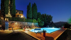 Une piscine avec des chaises longues, un mur de pierre et de grands arbres, le tout baigné par la lumière du crépuscule.