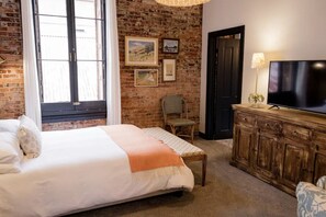 A bedroom with a brick wall, a bed with a white and orange blanket, a wicker chair, a wooden cabinet with a TV, and a door.