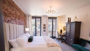 A bedroom with a brick wall, a bed with white bedding, a desk with a chair, and a dark cabinet.