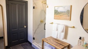 A bathroom with a dark door, a shower, a wooden vanity, a sink, a mirror, and a framed picture on the wall.