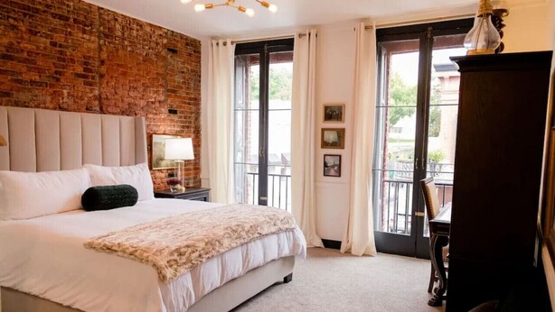 A bedroom with a brick wall, a large bed, a nightstand, a dresser, and a view through a window.