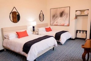 A hotel room with two beds, a nightstand, a lamp, a wall-mounted mirror, a large abstract painting, and a shelf with towels.
