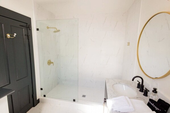 Une salle de bain dotée d’une douche en verre, d’un lavabo en marbre, d’un miroir avec un cadre doré et d’une porte en bois foncé.