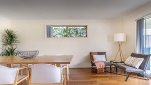 Una sala de estar moderna con una mesa de comedor de madera, sillas blancas, un sofá gris con cojines, una lámpara de pie y un amplio ventanal con vistas a árboles.