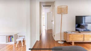 Una sala de estar moderna con piso de madera, un sofá blanco, una mesita pequeña blanca, una estantería, una lámpara de pie y un televisor sobre un mueble de madera.