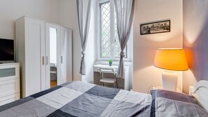 Quarto de hotel com cama, escrivaninha, cadeira, guarda-roupa, televisão e abajur.