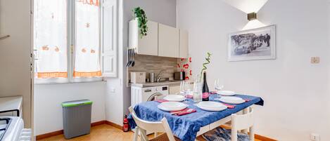 A dining area with a table set for two, a kitchen in the background, and a framed picture on the wall.