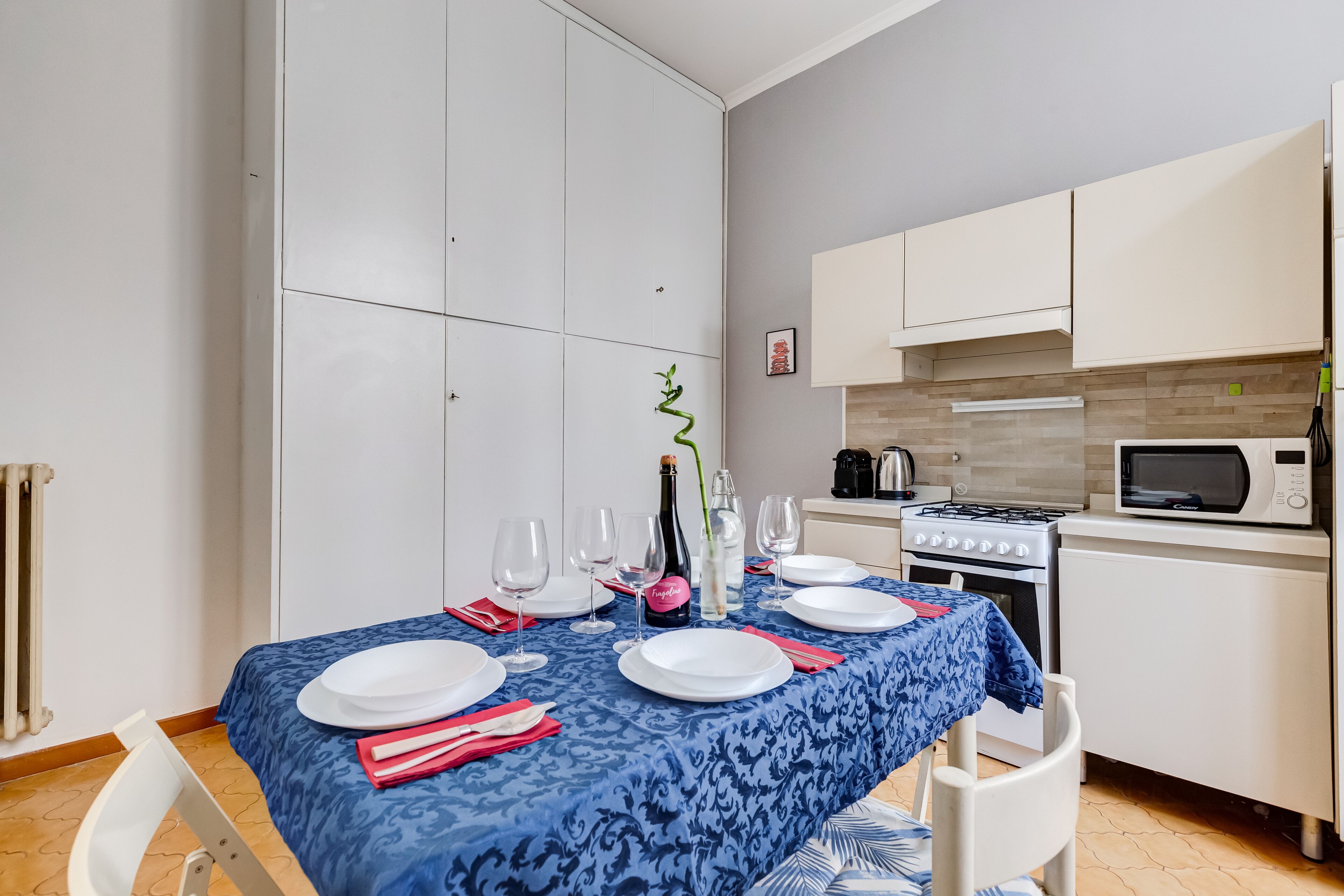 A dining area with a blue tablecloth, white plates, and a bottle of champagne. There is a kitchen with a microwave, stove, and cabinets in the background.