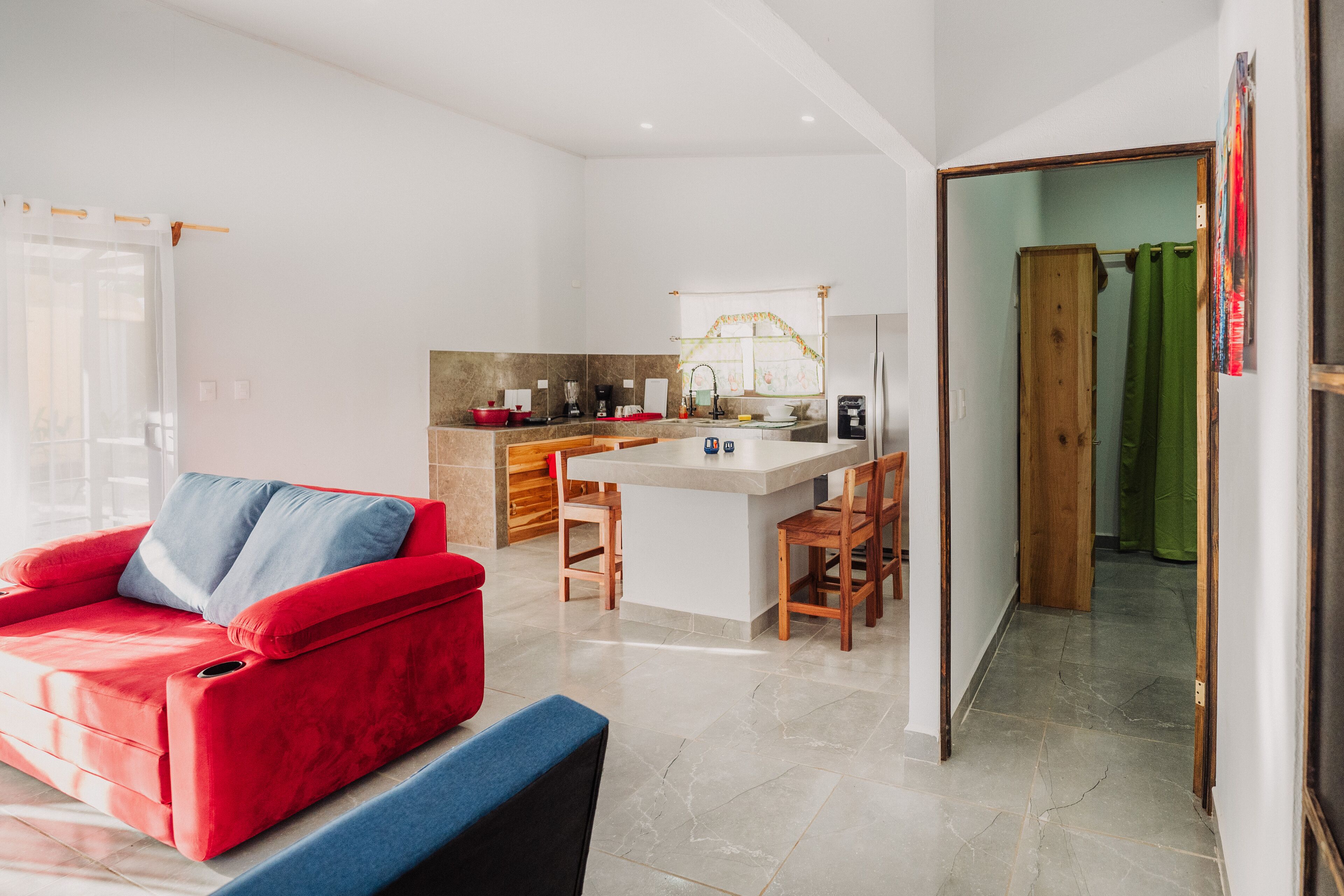 A modern living room with a red sofa, a kitchen area with a central island, and a hallway leading to a bathroom.