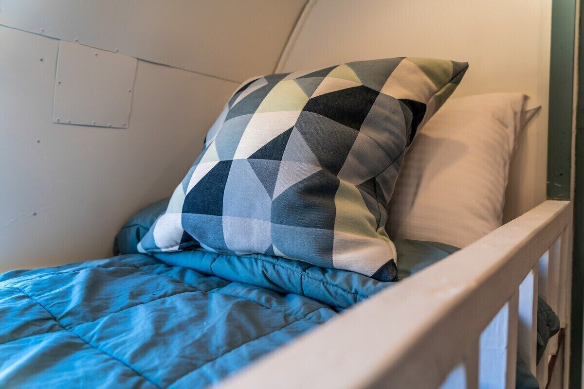 Ein Einzelbett mit einer blauen Bettdecke und einem gemusterten Kopfkissen in einem Zimmer mit weißer Wand und einem Holz-Kopfteil.