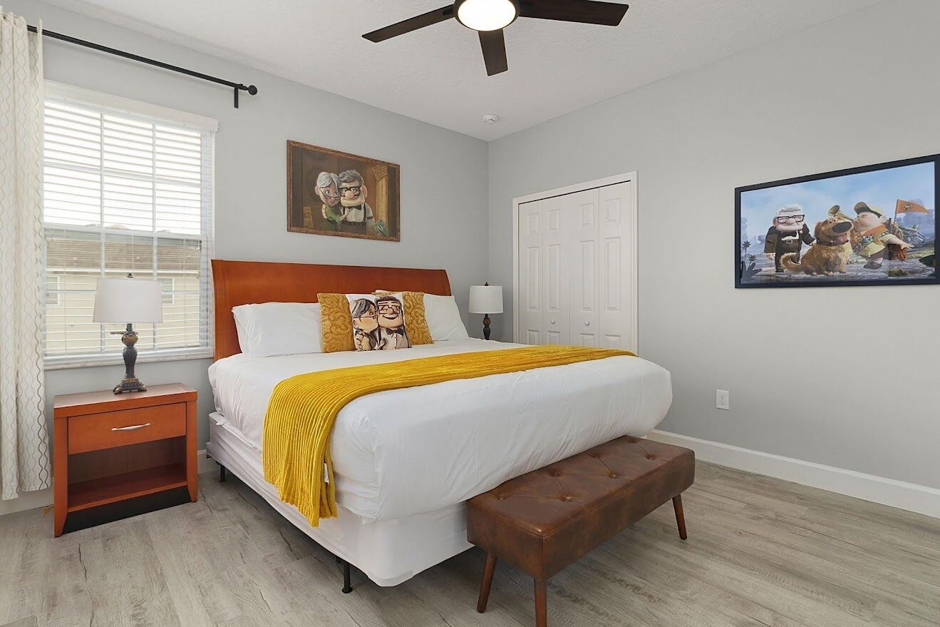 A bedroom with a bed, nightstand, bench, ceiling fan, window with blinds, and two wall-mounted pictures.