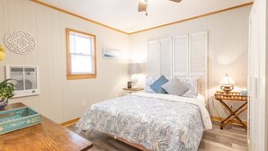 Ein Schlafzimmer mit Bett, Deckenventilator, Fenster, Nachttisch mit Lampe und Wandklimaanlage.
