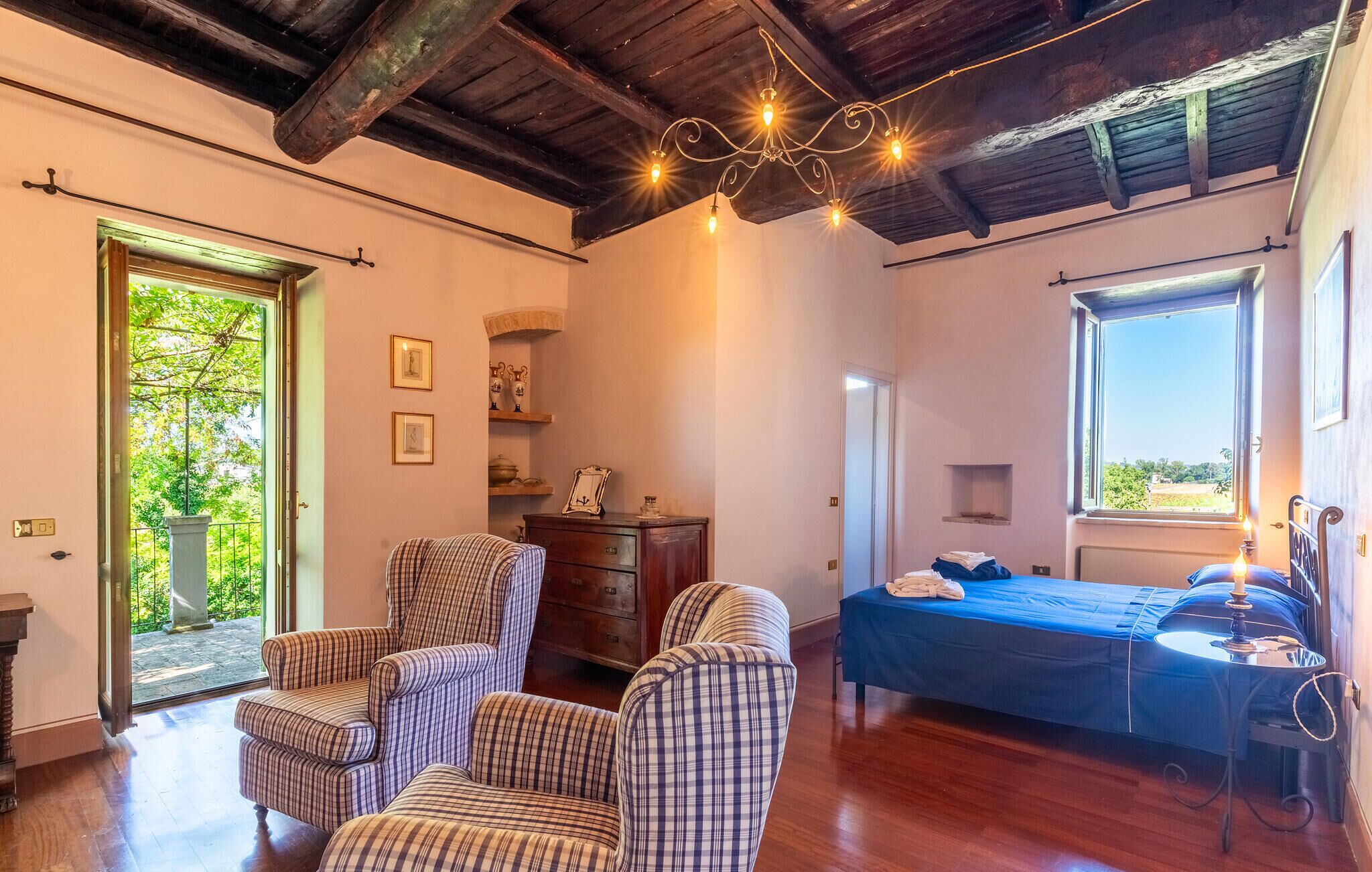 Une chambre avec un lit, deux fauteuils, une commode, un lustre et une fenêtre donnant sur un beau paysage.