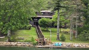 A lakeside cabin with a wooden staircase leading to a deck, surrounded by lush greenery and a stone wall.