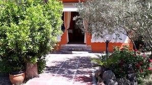 Un chemin mène à un bâtiment à la façade orange et au toit rouge. Des plantes en pot et un mur de pierre se trouvent sur la gauche, et un arbuste aux fleurs rouges sur la droite.