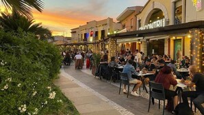 Un café animé en plein air, avec des tables et des chaises, entouré de verdure et de bâtiments dont les enseignes sont illuminées.