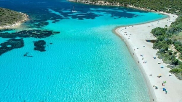 Vue aérienne d’une zone côtière avec une plage, des eaux turquoise et limpides, et un voilier.