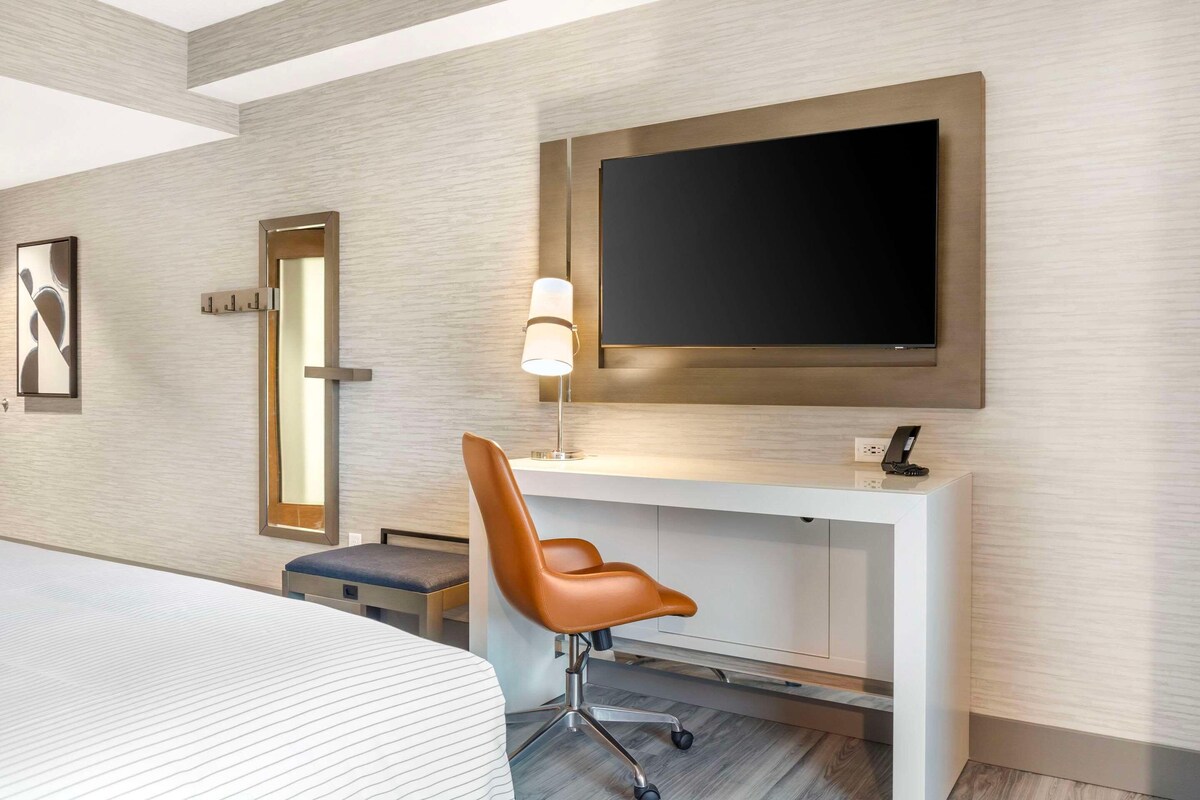 A modern hotel room with a large bed, a desk with a flat-screen TV, a chair, and a bedside table.