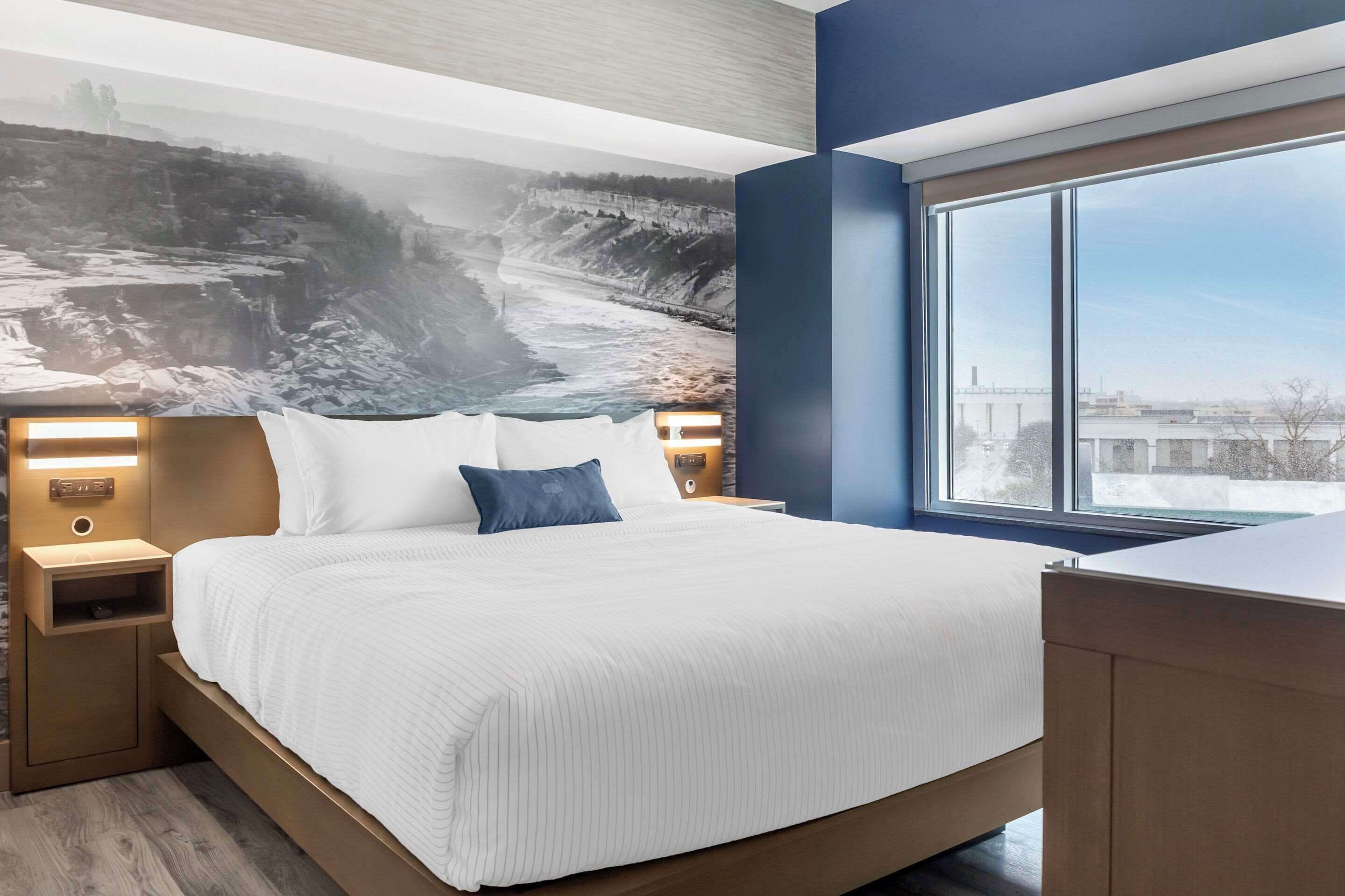Ett modernt hotellrum med en stor säng, ett sängbord, en vägglampa och ett stort fönster med utsikt över en bro och byggnader.