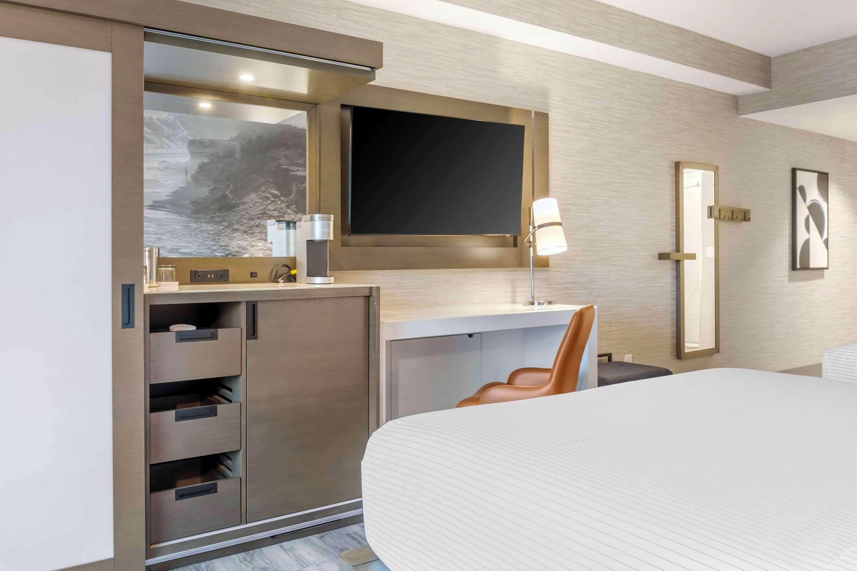 A modern hotel room with a large bed, a desk with a chair, a flat-screen TV, a mirror, and a wardrobe with multiple drawers.