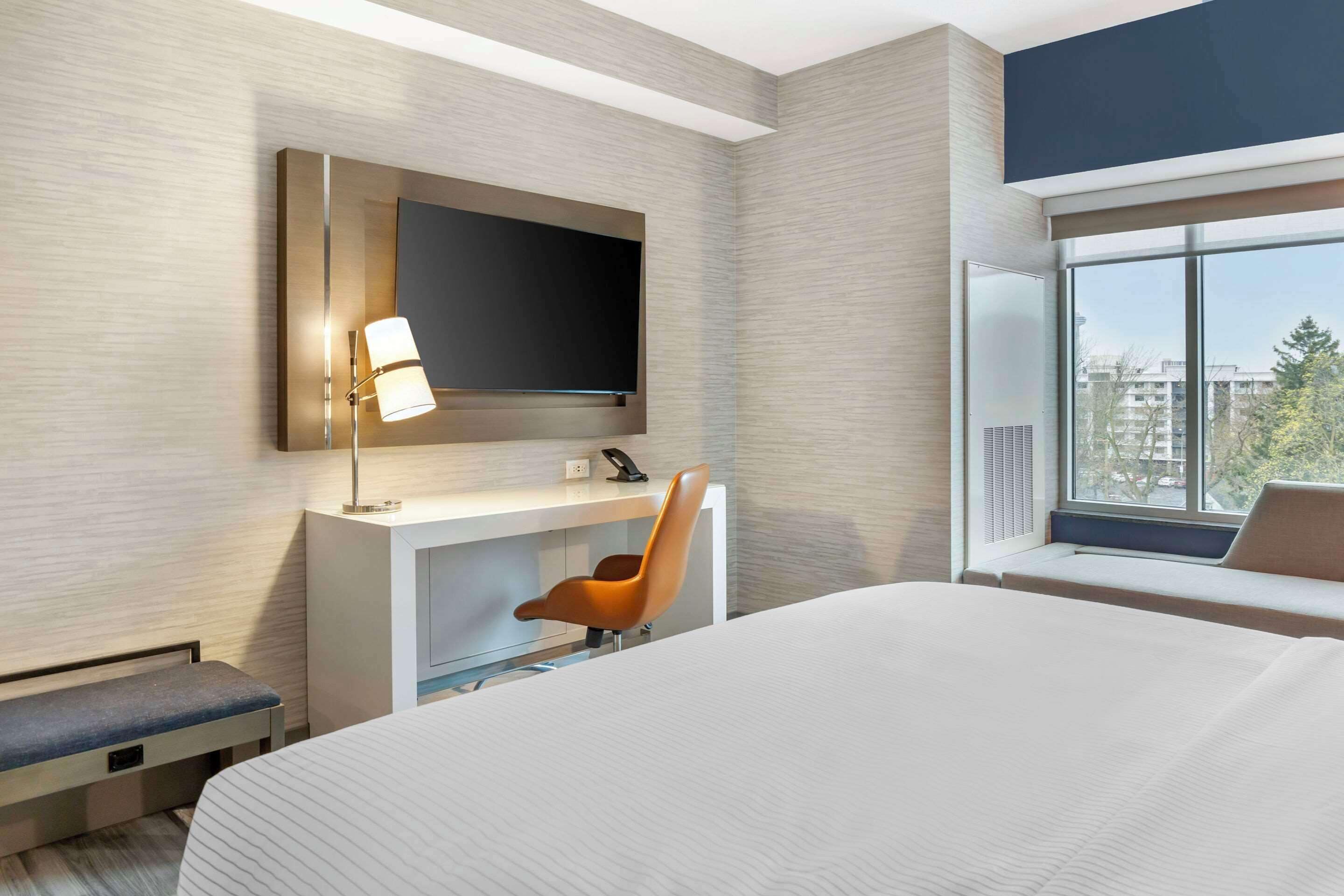 A modern hotel room with a large bed, a desk with a chair, a flat-screen TV mounted on the wall, and a window with a view of buildings and trees.