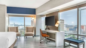 Une chambre d’hôtel comprenant un lit, deux fauteuils, un bureau avec un ordinateur, une télévision et de grandes fenêtres donnant sur la ville.
