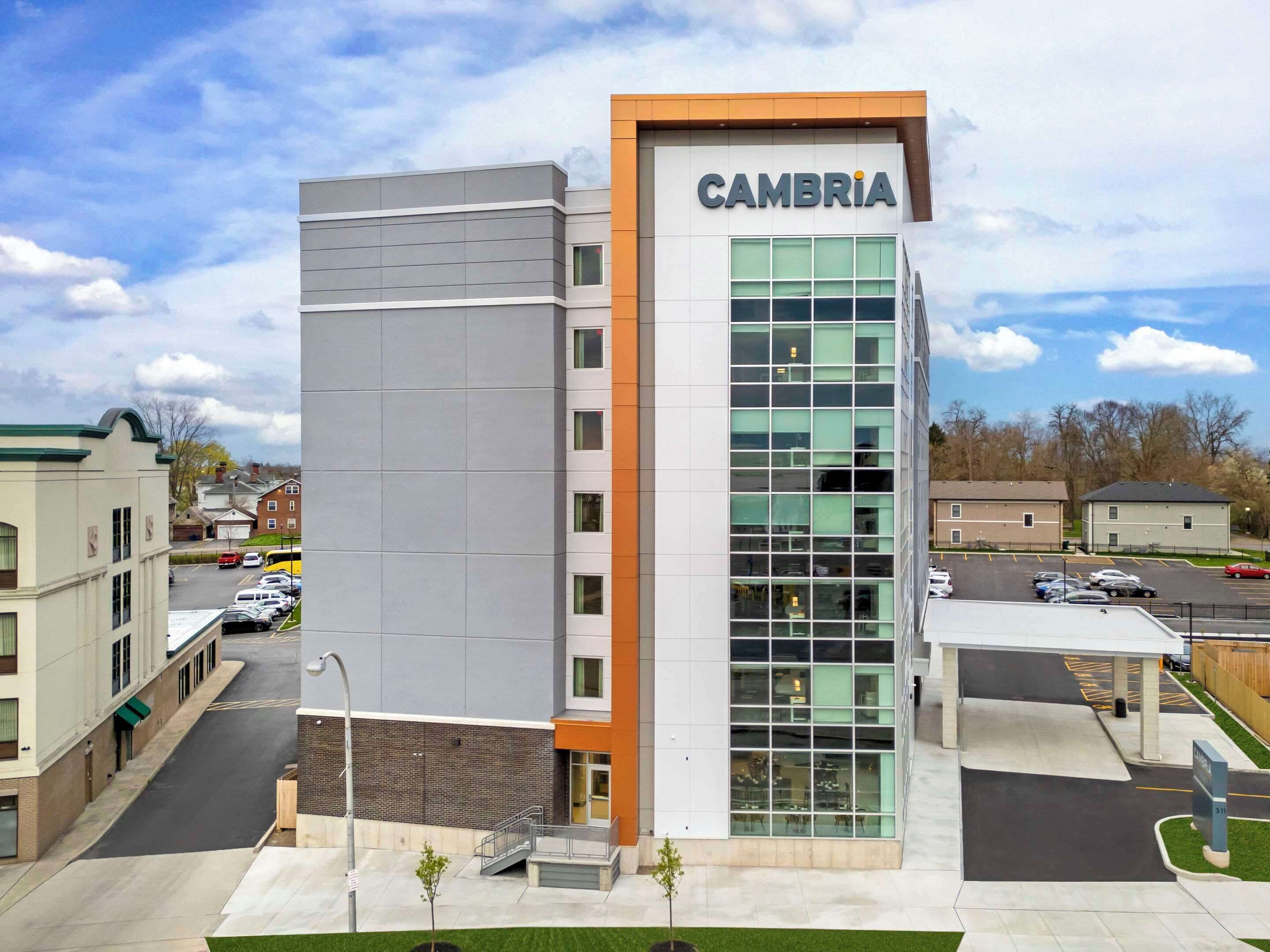 A modern building with a prominent 'CAMBRIA' sign, featuring a mix of glass and solid facade, and a parking area in front.