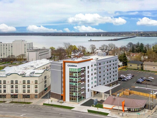 A modern hotel with a prominent 'Cambria' sign, a parking lot, and a view of a body of water with a distant island.