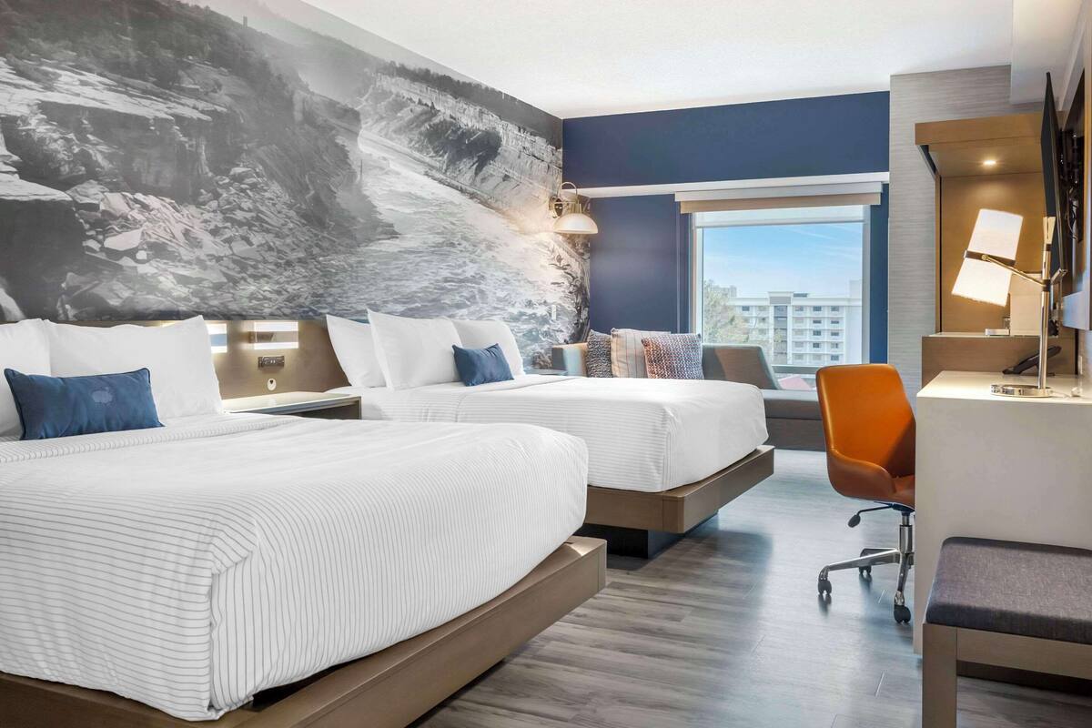 Ein Hotelzimmer mit zwei Betten, einem Schreibtisch, einem Stuhl und einem großen Fenster mit Blick auf die Stadt.