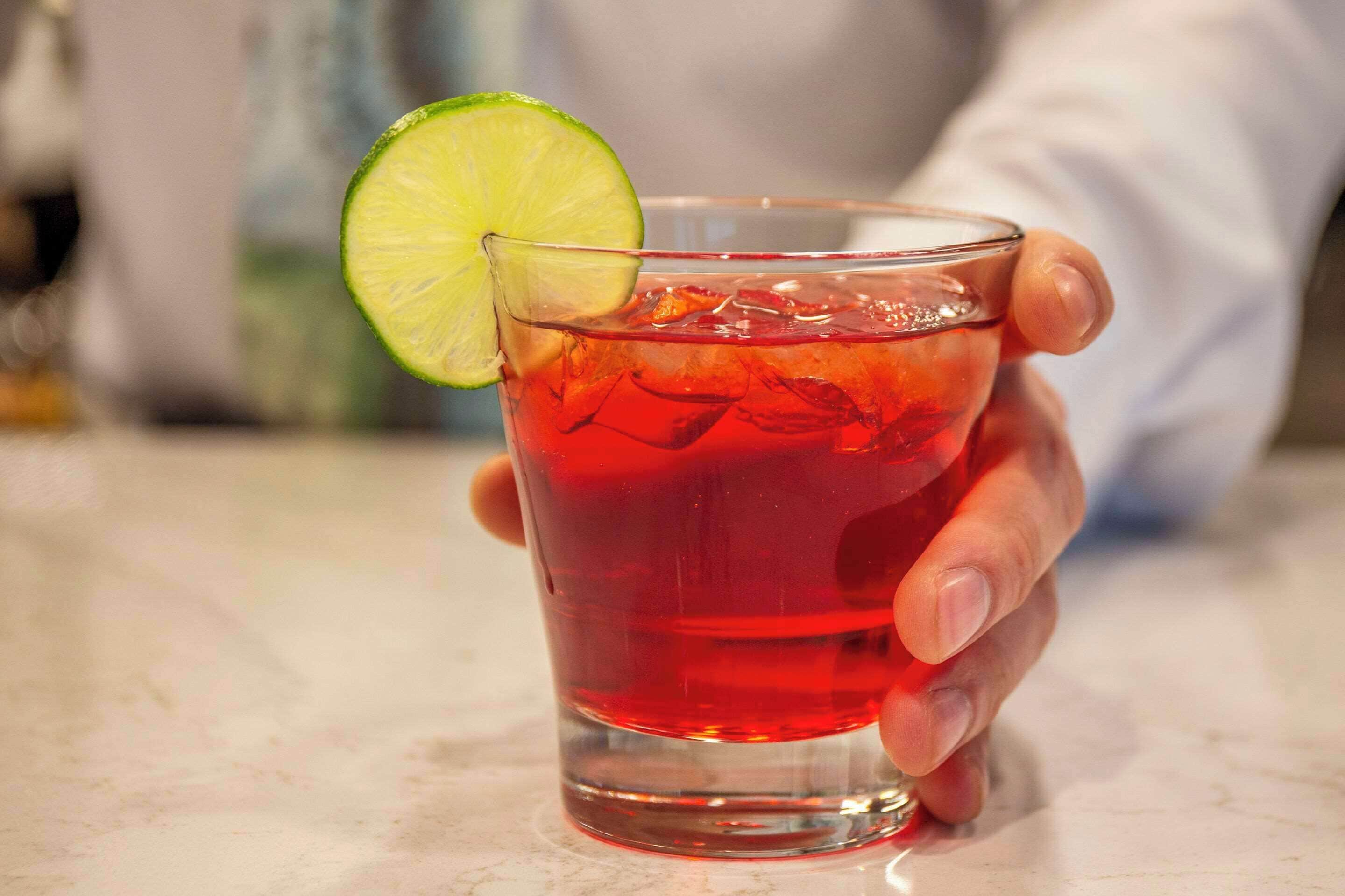 Una mano sosteniendo un vaso con una bebida roja y una rodaja de lima.