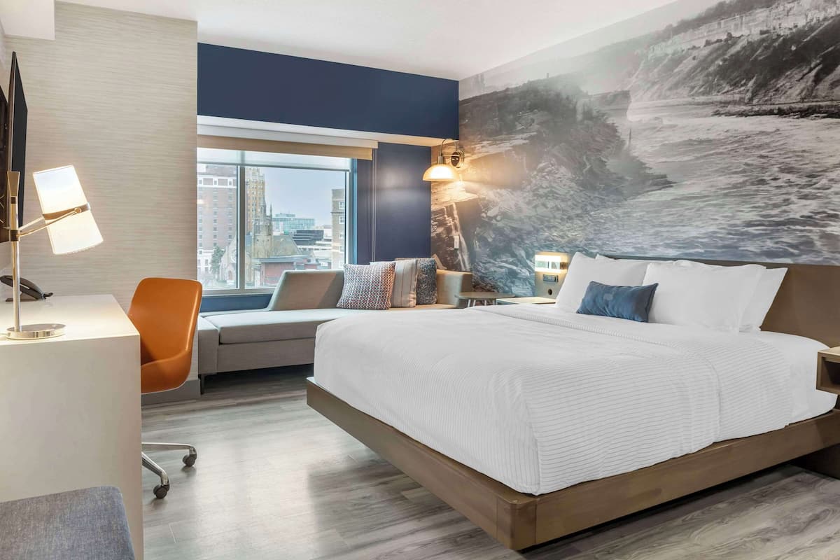 Ein modernes Hotelzimmer mit einem großen Bett, einem Schreibtisch und einem Stuhl. Ein großes Fenster bietet Blick auf die Stadt, an der Wand hängt ein Gemälde.
