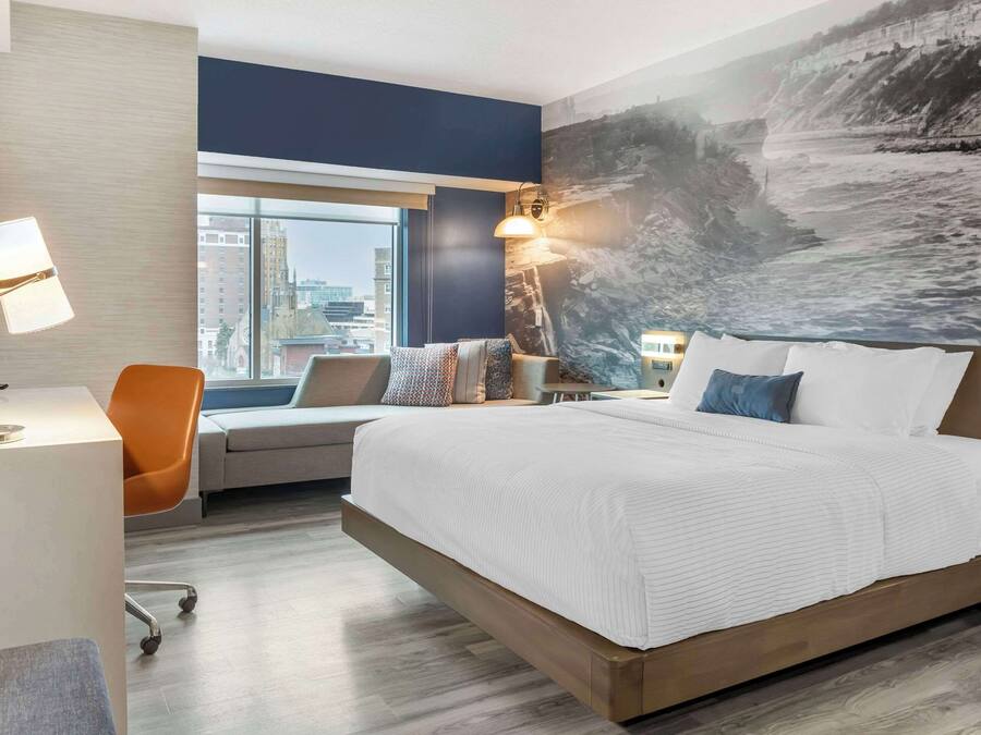 A modern hotel room with a large bed, a desk, and a chair. There is a large window with a city view and a painting on the wall.