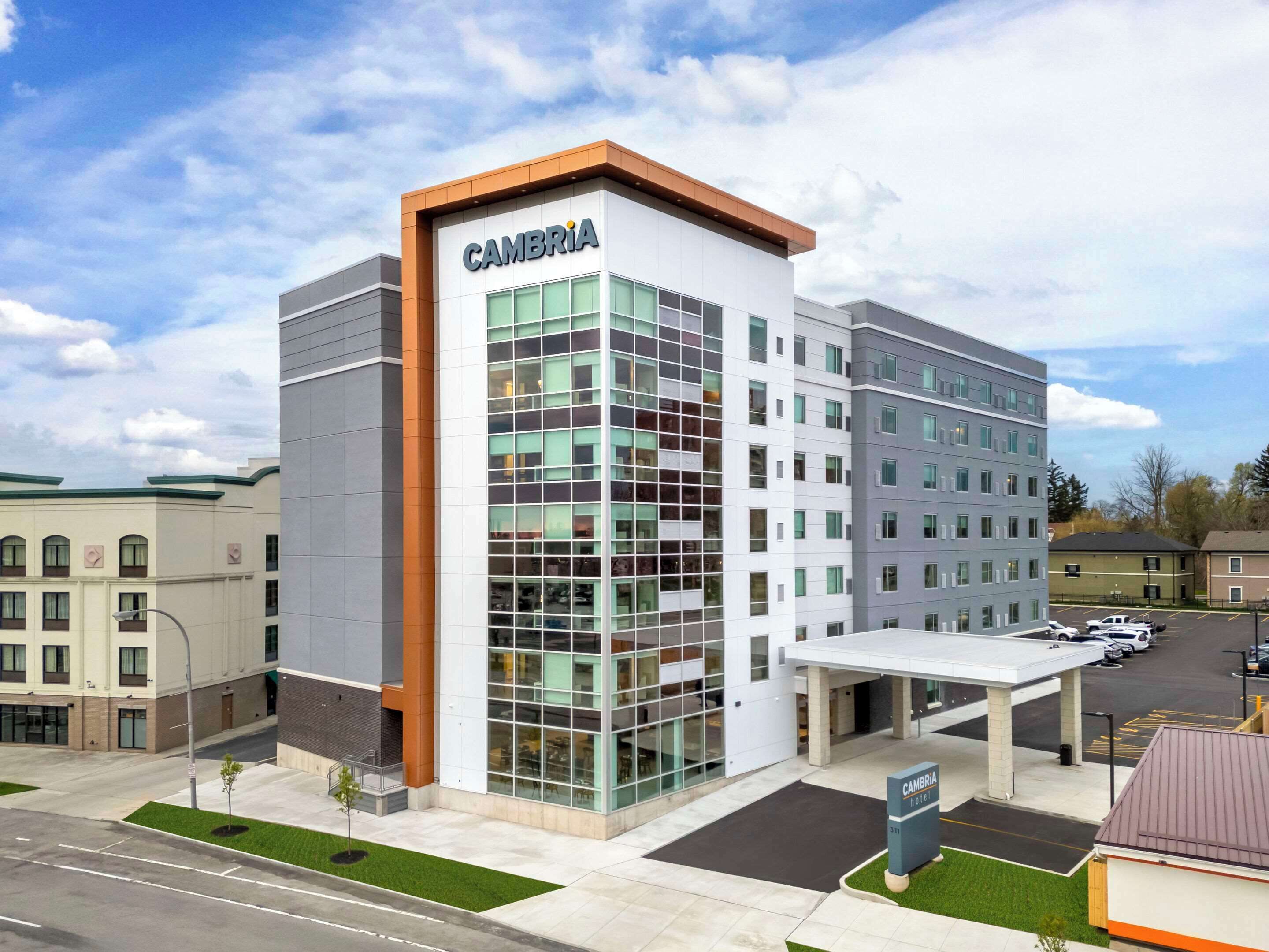 Un hôtel moderne, reconnaissable à son grand panneau « CAMBRIA », doté d’une façade en verre et d’un espace de stationnement situé à l’avant.