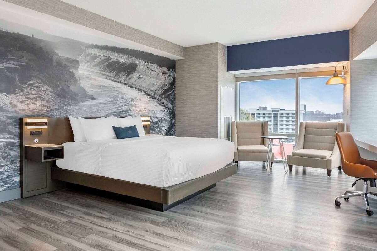 Ein modernes Hotelzimmer mit einem großen Bett, einem Schreibtisch, zwei Stühlen und Blick auf die Stadt durch große Fenster.